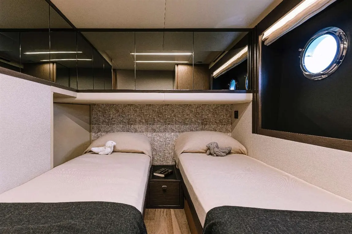 Modern cabin with two single beds, soft linens, and decorative toys, featuring a round window and mirrored storage.